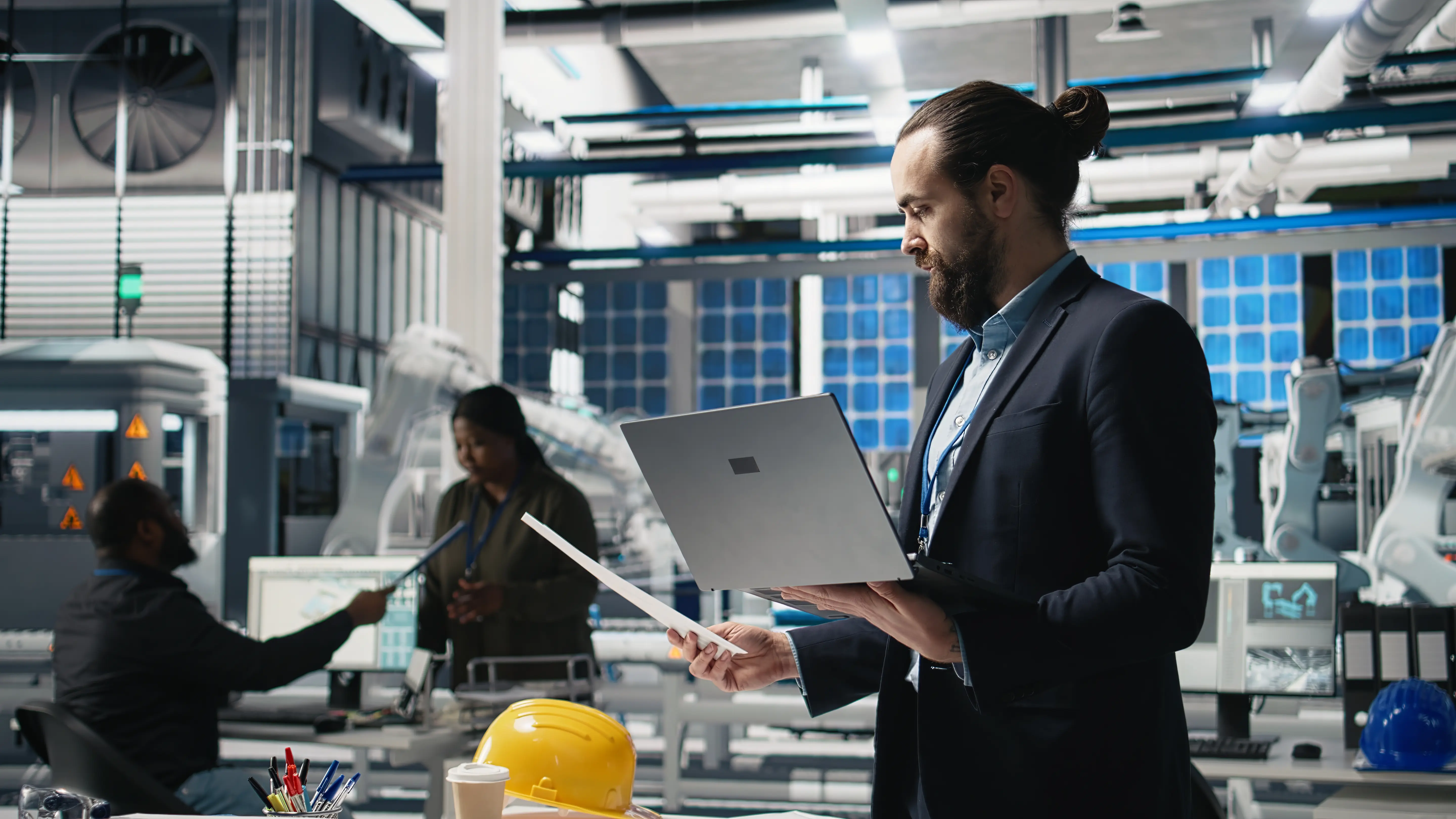 AI Business Automation. Professional in an industrial workspace using a laptop while reviewing operations, with another team member working in the background.