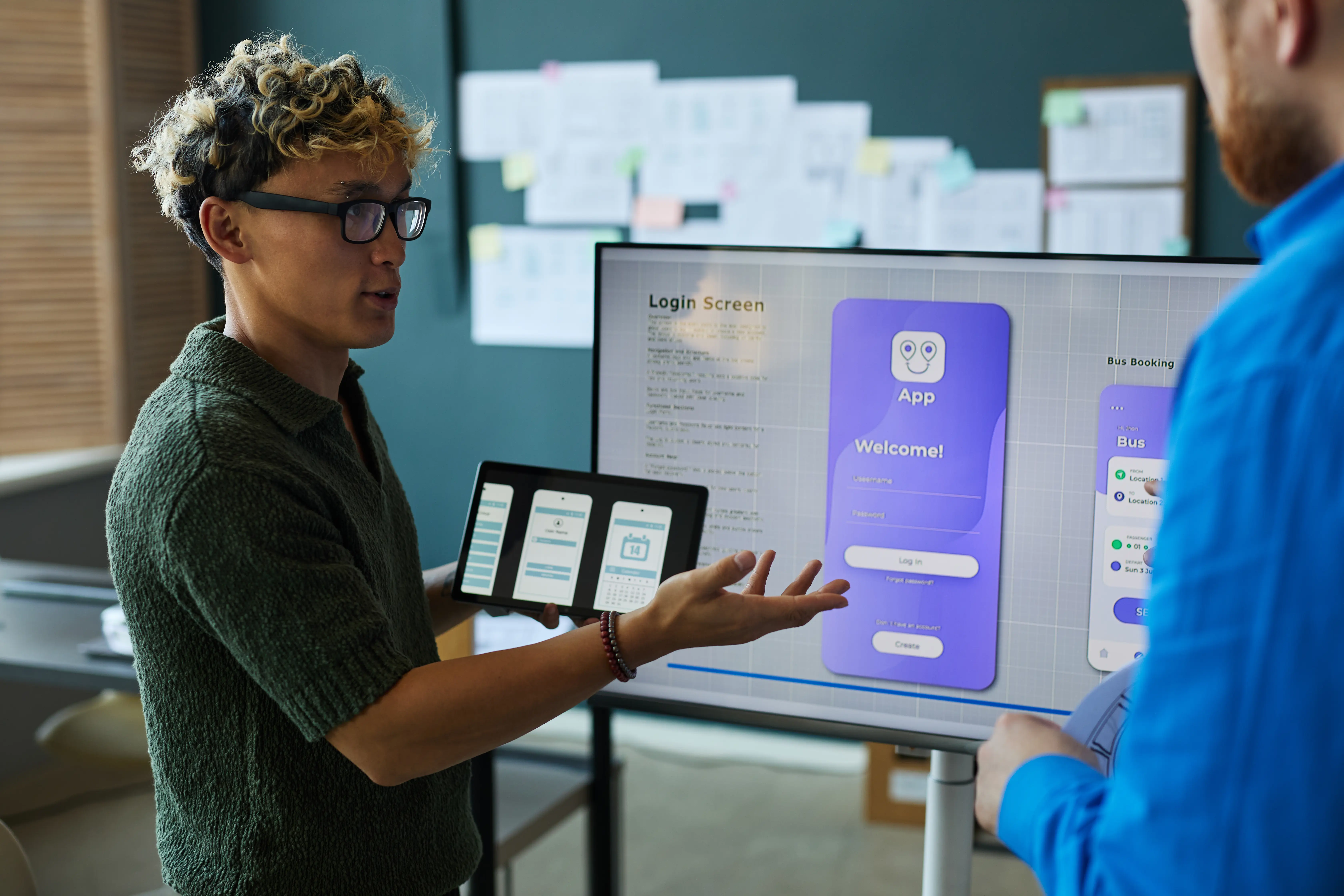 A product designer presenting a mobile application prototype on a large display while discussing user interface elements and functionality with a colleague.